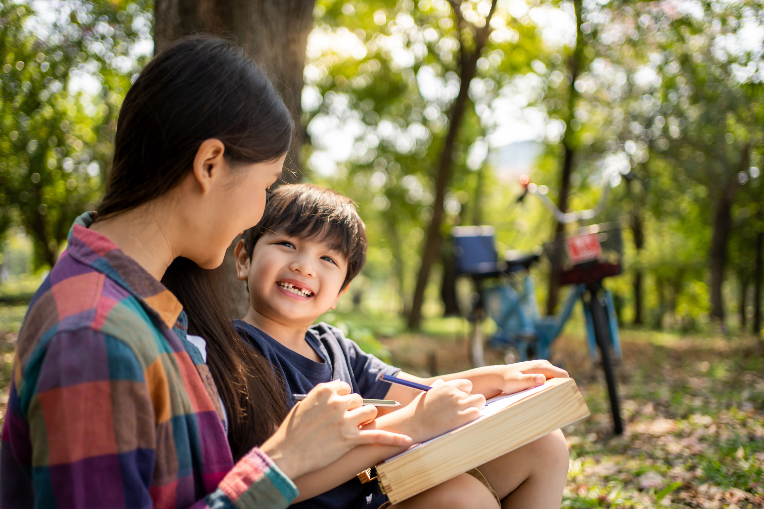 An East Asian mother and young child paint together in the woods. Behind them is their bicycle.