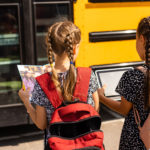 The back of two young female presenting children stand waiting for a schoolbus. One looks at a book, one looks at a tablet.