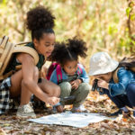 Three young kids crouch in the woods around a scavenger hunt map. One looks at the map with binoculars.